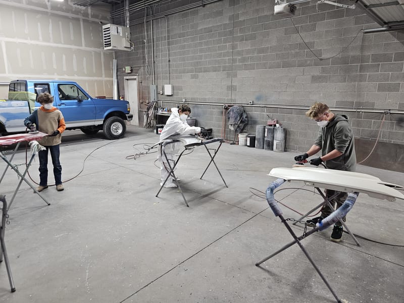 Three technicians sanding separate body panels on stands in the workshop