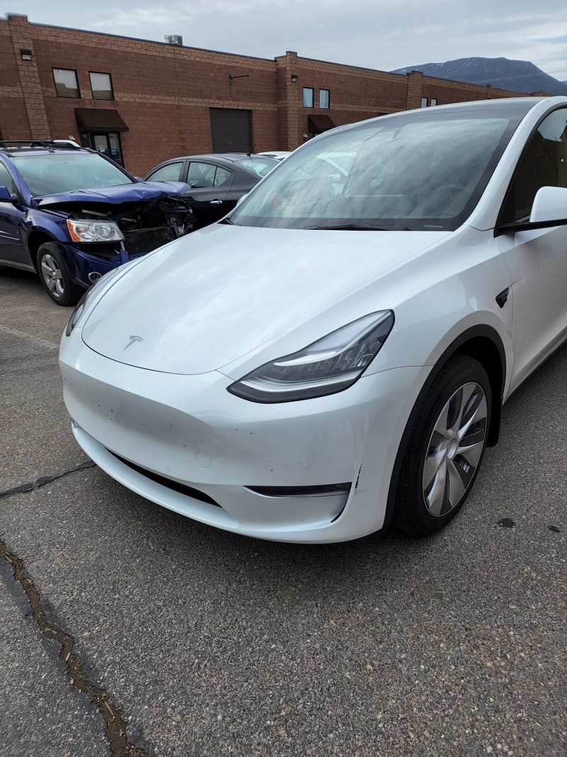 The white Model Y parked on the shop lot, a heavily damaged blue SUV behind it