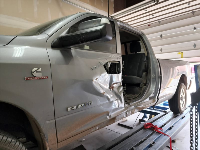 A silver Ram 3500 crew cab clamped to the frame rack, its driver's door torn open and the cab side pushed in